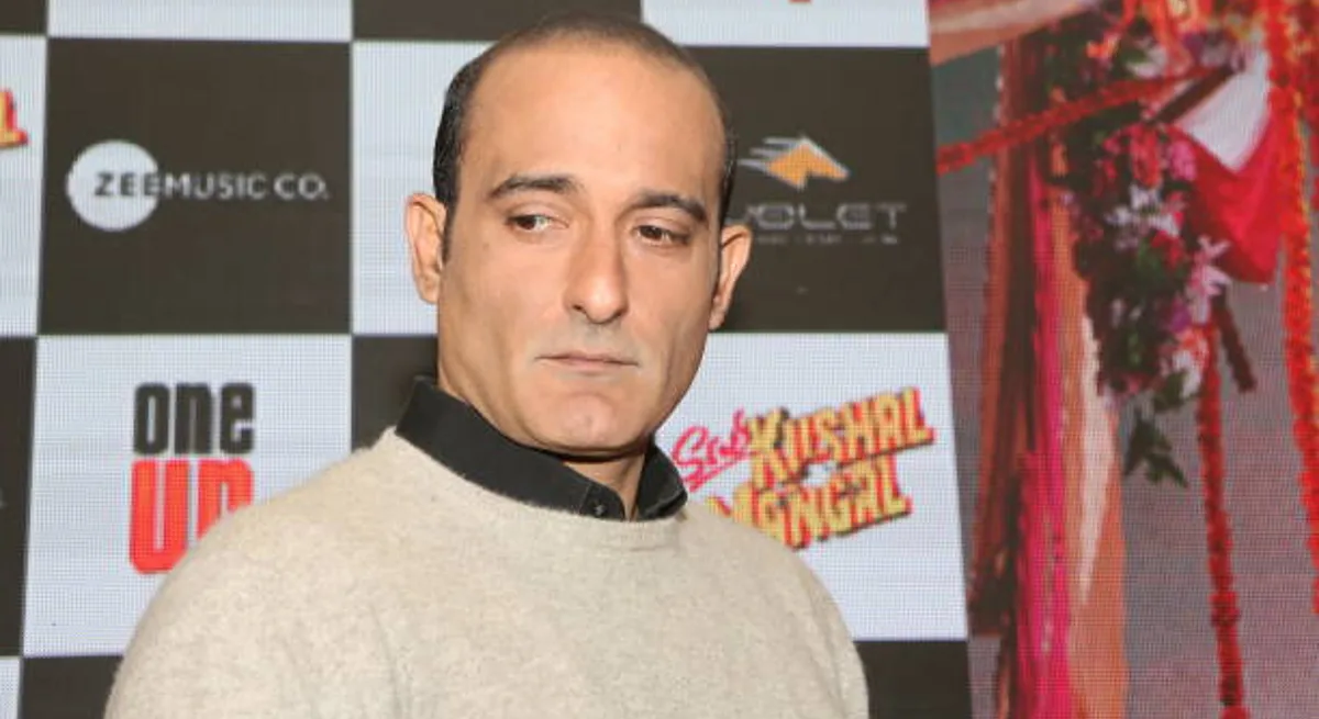 Akshaye Khanna at a film promotion event, cream sweater over a dark collar, against a branded backdrop