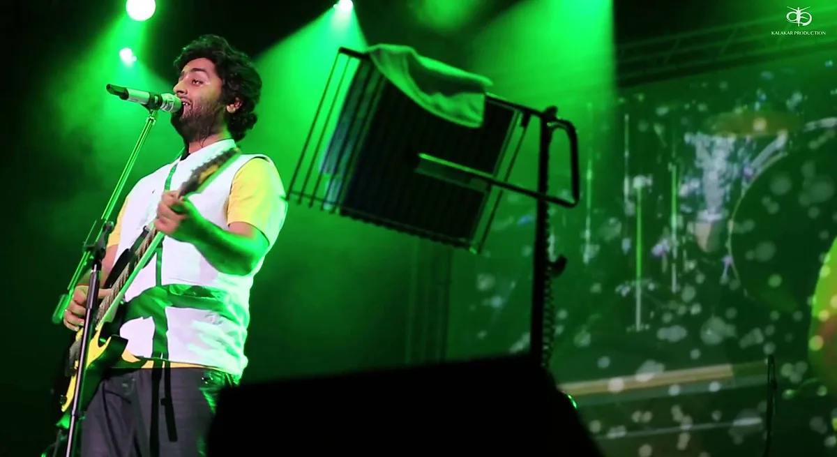 Arijit Singh singing and playing guitar on a green-lit concert stage