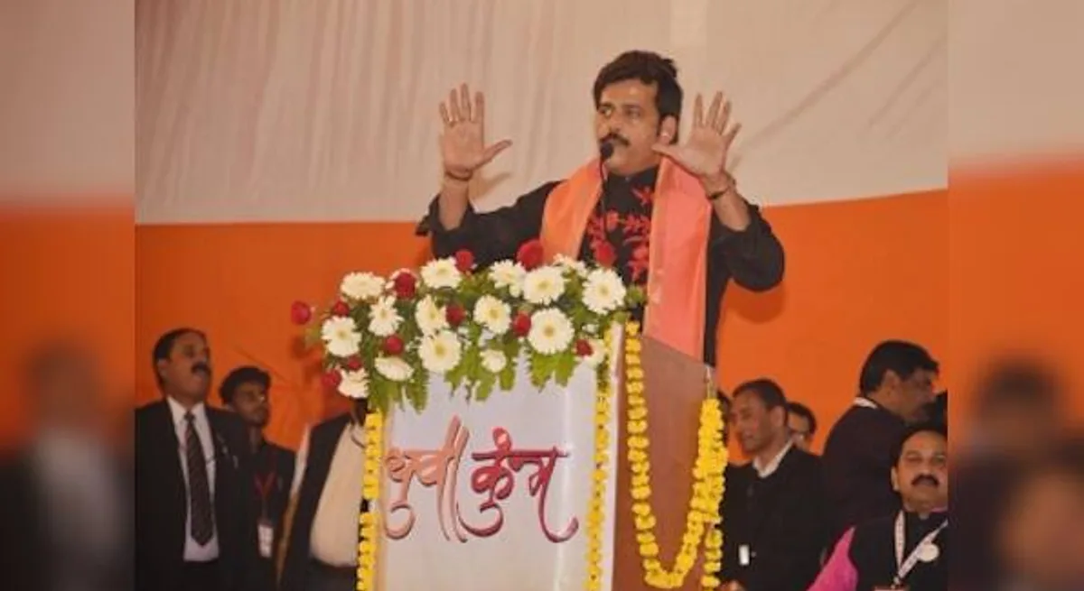 Ravi Kishan addressing a BJP public meeting from a flower-decked podium, both hands raised, saffron scarf around his neck