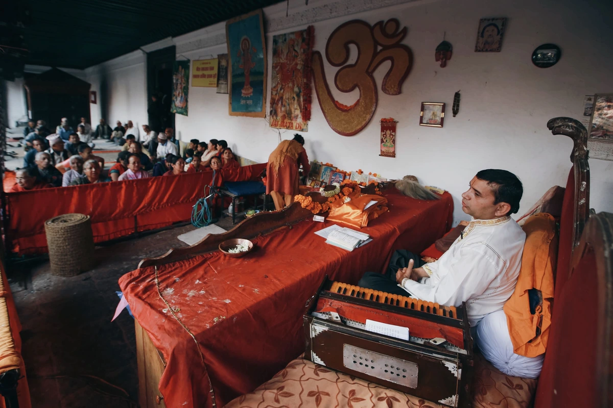 A vocalist seated at a harmonium during a devotional Indian classical performance, with an audience listening in a hall