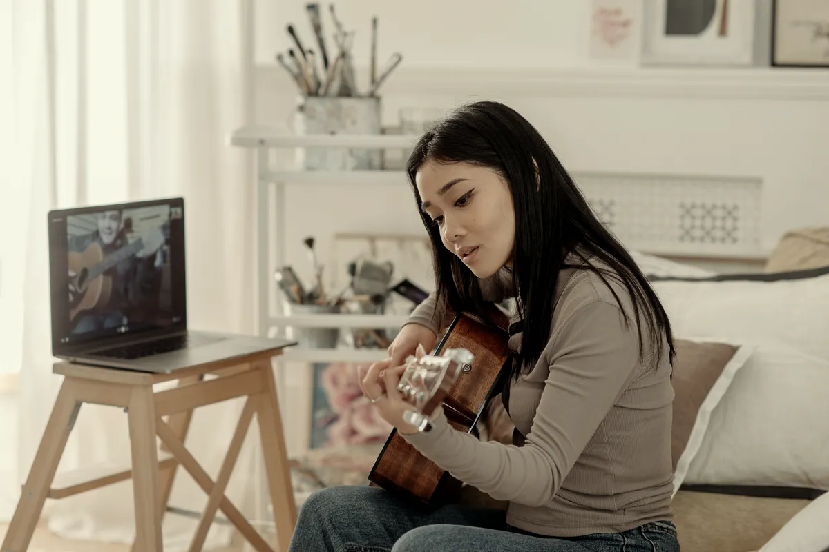 A student following an online music lesson on a laptop while playing a ukulele at home