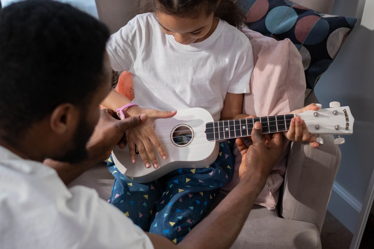 A young girl holding a white soprano ukulele while an adult guides her fingers on the fretboard